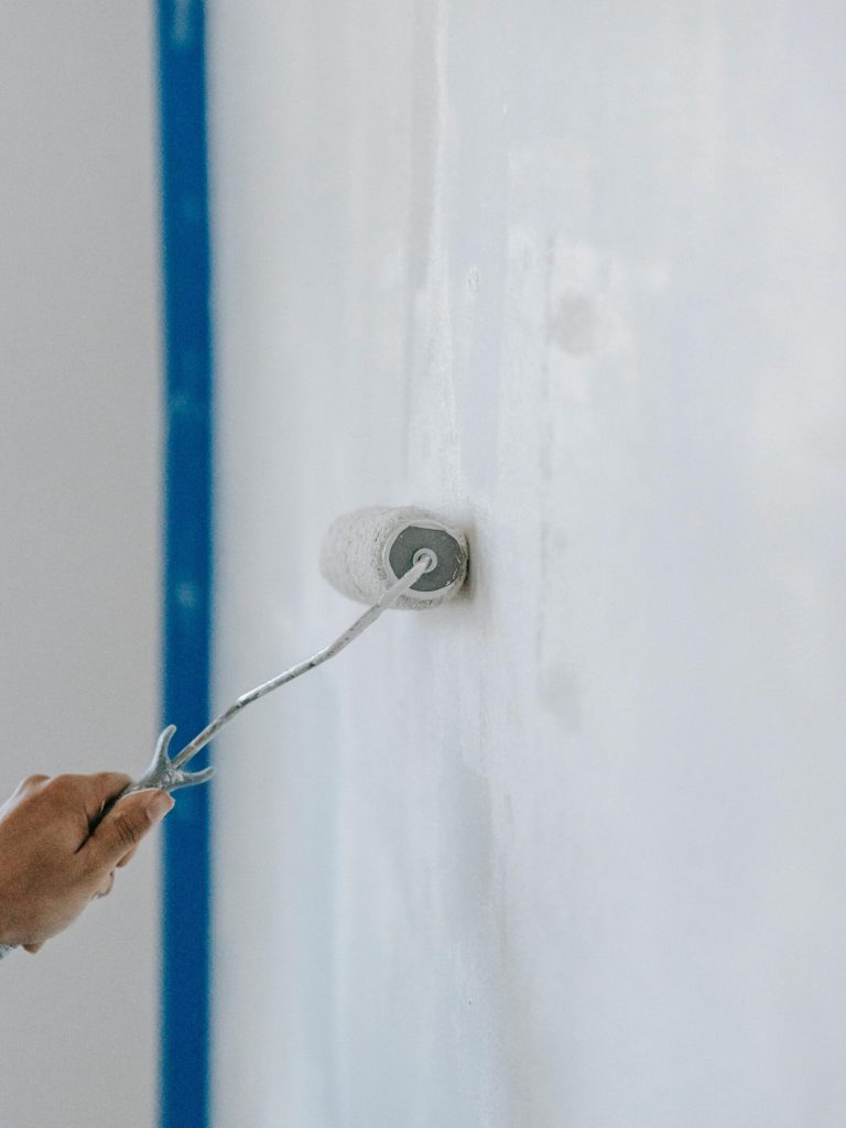 A close-up of a person using a paint roller to apply white paint on an indoor wall.